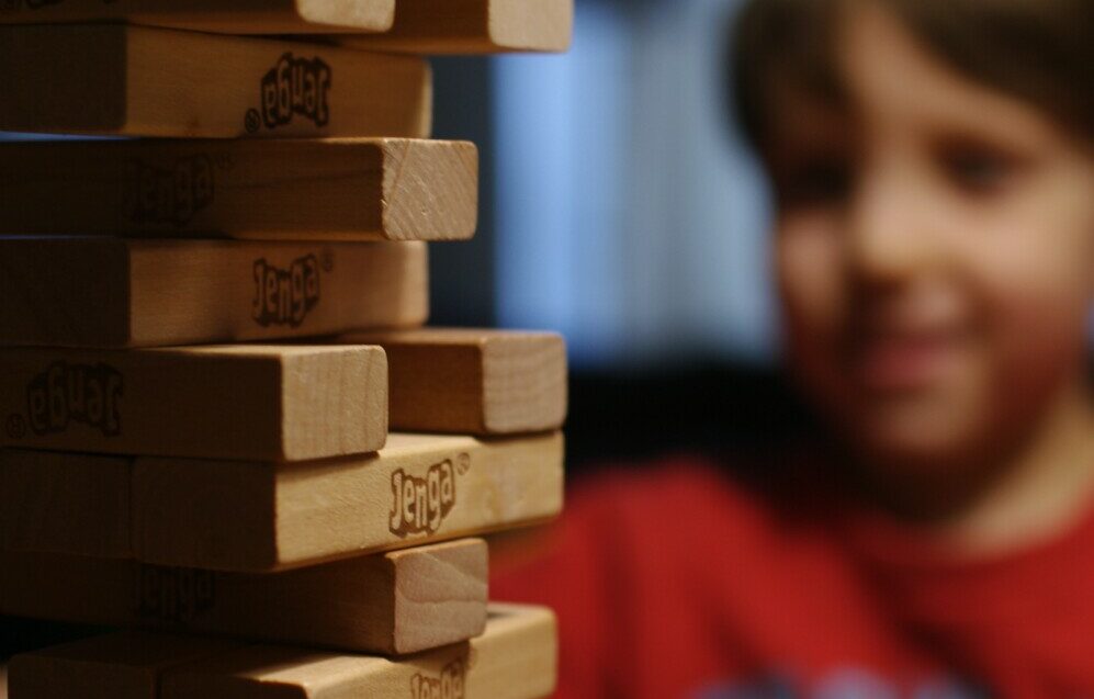 Ein Jenga-Turm aus Holz ist zu sehen, im Hintergrund verschwommen ein Kind mit rotem Pullover