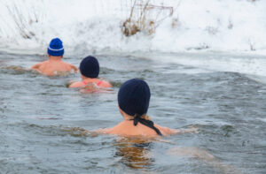 Drei Personen die hintereinander in einem wirtlichen See schwimmen. Die Personen tragen Mützen. Der See ist umgeben von Schnee.