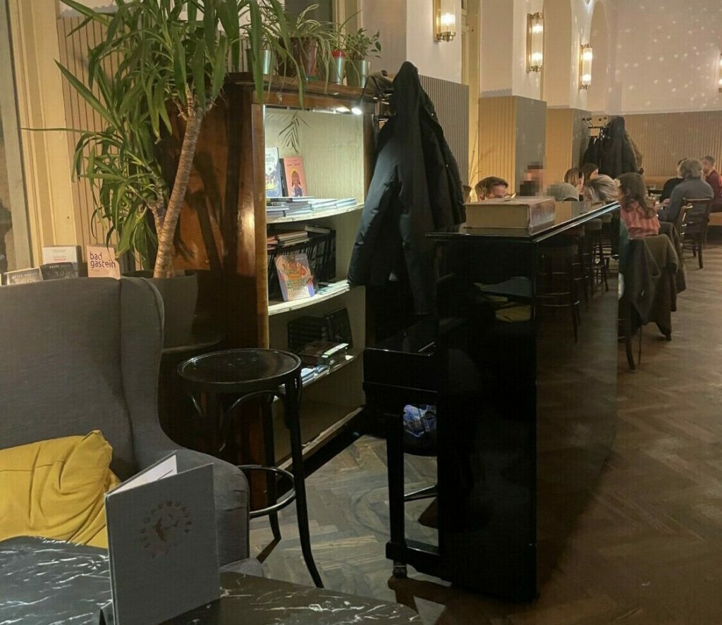 Ein Foto des Cafe Schopenhauers, in dem ein schwarzes Wandklavier, ein kleines Bücherregal und sitzende Menschen zu sehen sind.